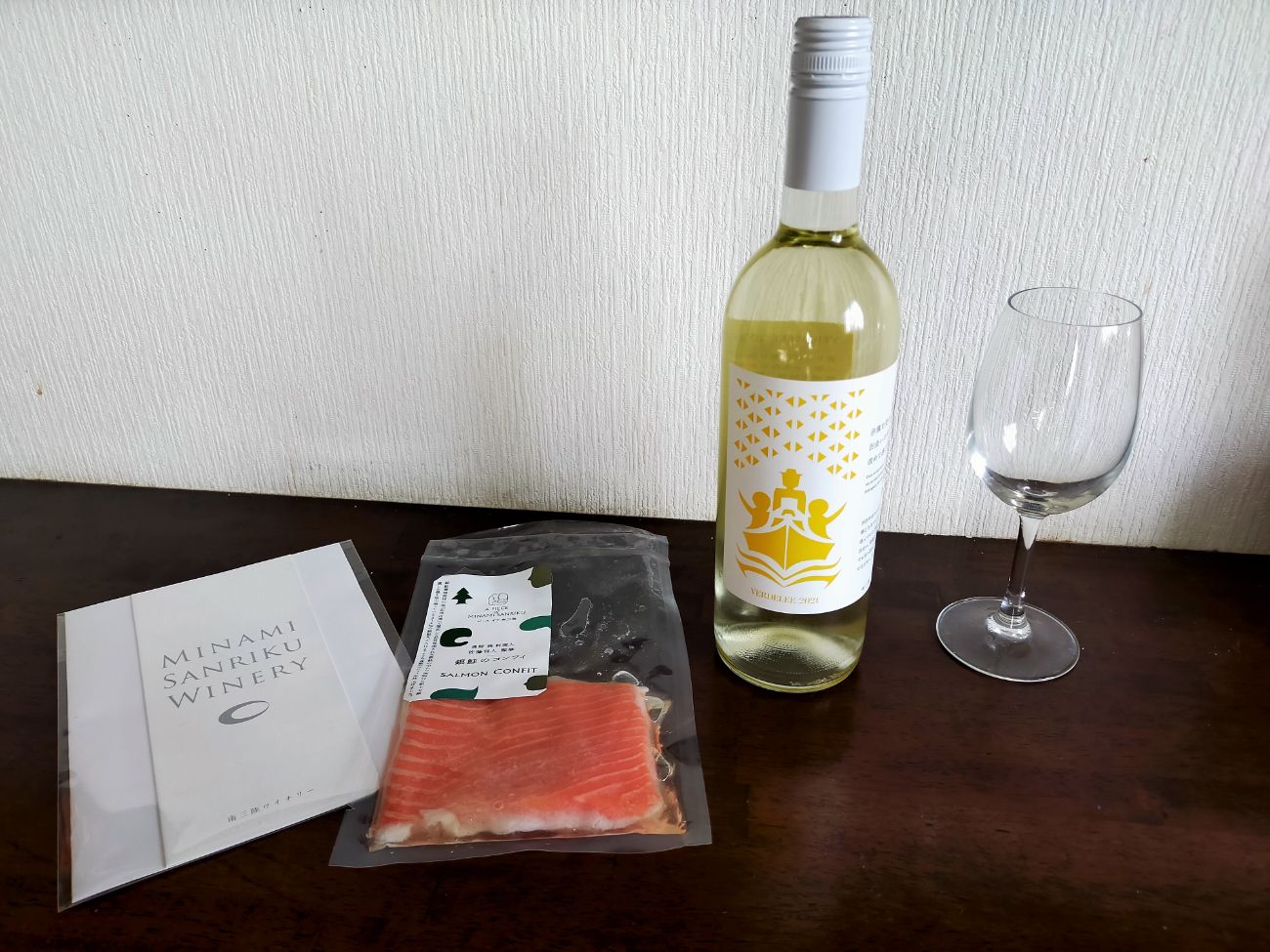 「MINAMI SANRIKU WINERY」と書かれたパンフレット、真空パックされたサーモン、白ワインのボトル、ワイングラスが木のテーブルの上に並んでいる写真