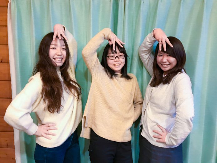 水色のカーテンの前に並んで立った3人の女性が、笑顔で頭の上に3本指を出した手を乗せたポーズをしている写真