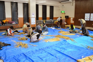 多くの参加者が青いシートの上に座り、グループで藁を使ってしめ縄作りをしている会場の全体写真