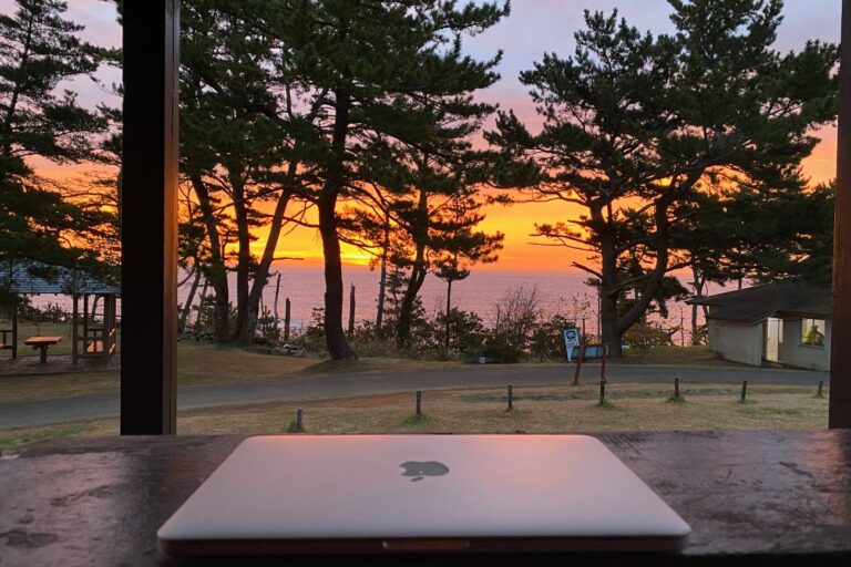 夕焼けの空と海を背景に、木々が立ち並ぶ静かな公園のベンチの上にノートパソコンが置かれている写真