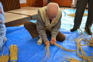 高齢男性が青いシートの上に座り、手で藁をまとめてしめ縄の一部を編んでいる作業中の写真