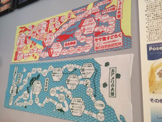 黄色と赤、水色を基調に配色された「サケ生すごろく手ぬぐい」と、水色と白、緑で配色された「カメ生すごろく手ぬぐい」が上下に並んで壁に掲示されている写真