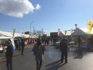 左右にのぼりを立てた白い屋根のテントが立ち並び、その間を来場者たちが歩いている写真