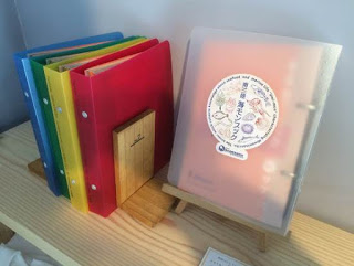 木製の棚の上に、赤・黄・緑・青のカラフルなリングファイルが縦に並べられ、その横には魚などのイラストがカラフルに描かれた丸いシールが貼られた透明のカバー付き冊子が立てかけられて展示されている様子の写真