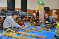 複数の高齢者が青いシートの上で藁を束ねてしめ縄を編む作業をしている様子の写真