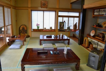 日差しの入る畳の客間に木製の座卓が整然と配置され、壁際に飾られた置物や額縁が置かれている部屋の写真