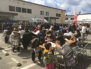 屋外にテーブルと椅子を並べて設けられた飲食スペースが、家族連れなどで満席になっている様子の写真