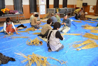 多くの参加者が青いシートの上で藁を使ってしめ縄作りをしている様子の別角度の写真