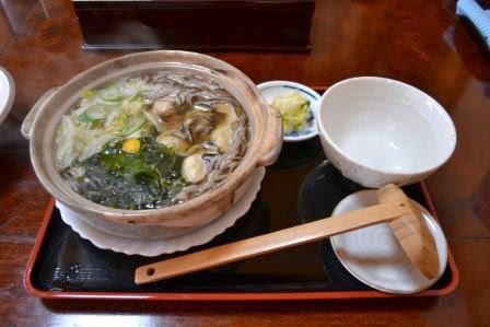 湯気の立つ土鍋にそばと牡蠣、野菜などが盛られたあつあつの牡蠣そばが黒いお盆にのせられている写真
