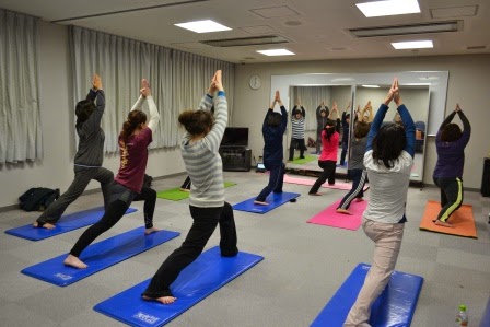 部屋の前方の大きな鏡の前で数名の女性たちがヨガマットの上で足を前後に開き、腕を頭上で合わせた同じポーズをとっているストレッチの教室の様子を写した写真