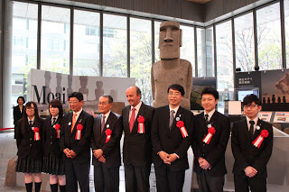 モアイ像の前にスーツ姿の関係者や制服姿の高校生が整列して笑顔で記念撮影をしている写真