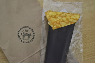 自然卵農園のロゴが入った紙袋の隣に黒い包みに包まれた三角形のクレープが置かれている写真