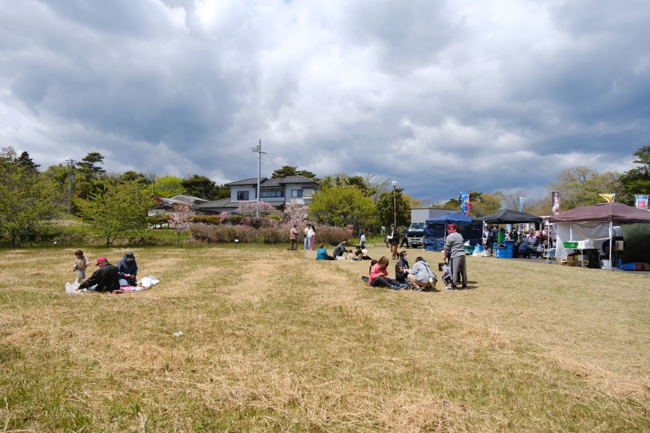分厚い雲が広がったり空の下、テントが並んだイベント会場の裏手にある芝生広場の一角で、複数の家族連れのグループがレジャーシートを敷いて座りのんびりくつろいでいる様子の写真