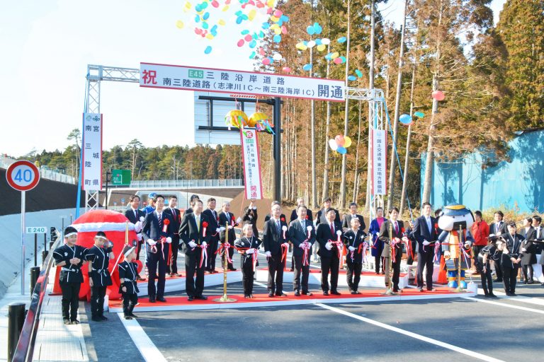 上空に風船が舞い上がり「南三陸道路開通」と書かれた横断幕が掲げられ、レッドカーペットの上に胸章リボンをつけた関係者や子供が横一列に習い、テープカットを行っている様子の写真