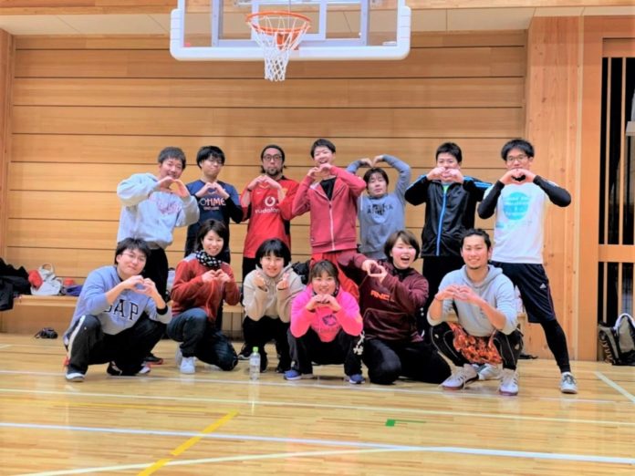 体育館のバスケットゴールの前で、ユニフォームや運動着を着た13人の人物が2列に並んでポーズを取っている集合写真