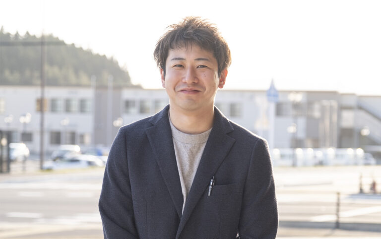 遠くの建物をバックに、紺色のジャケットを着た佐藤豪さんをアップで撮影した写真