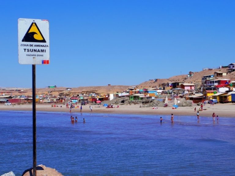 海岸沿いに「ZONA DE AMENAZA TSUNAMI HAZARD ZONE」と書かれた津波警戒区域の標識が立っており、背景には広がる砂浜と、赤や青などのカラフルな建物が並ぶ町並みが見える写真