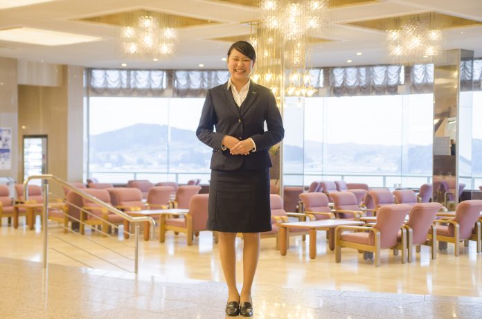 たくさんのテーブルセットが並ぶ広いロビーでスーツ姿の沖澤鈴夏さんが笑顔で立っている写真