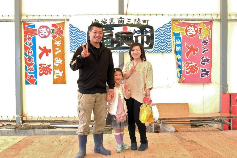 2枚の大漁旗と福興市の横断幕で飾られた壁の前で工藤さん夫婦と光奈乃ちゃんが並んでピースをしている写真