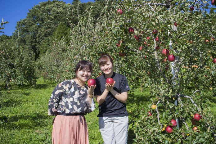 りんご畑で赤いりんごを手に笑顔で並ぶ女性リポーターと大沼さんの写真