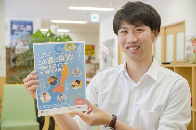 大きなポスターが貼られた室内で、白いシャツを着た男性が、笑顔で一枚のチラシカメラに見せている写真