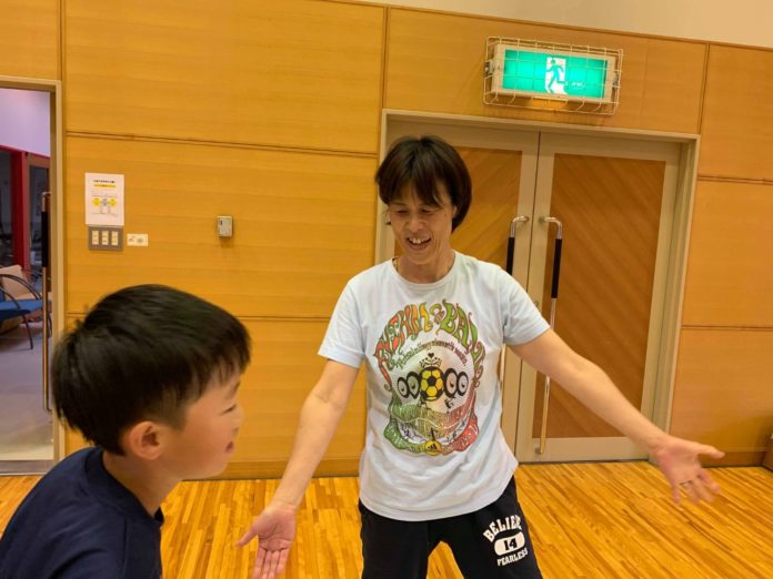 避難口誘導灯が設置された体育館の扉の前で、Tシャツ姿の女性が明るい笑顔を浮かべながら腕を広げて子どもに話しかけるている写真