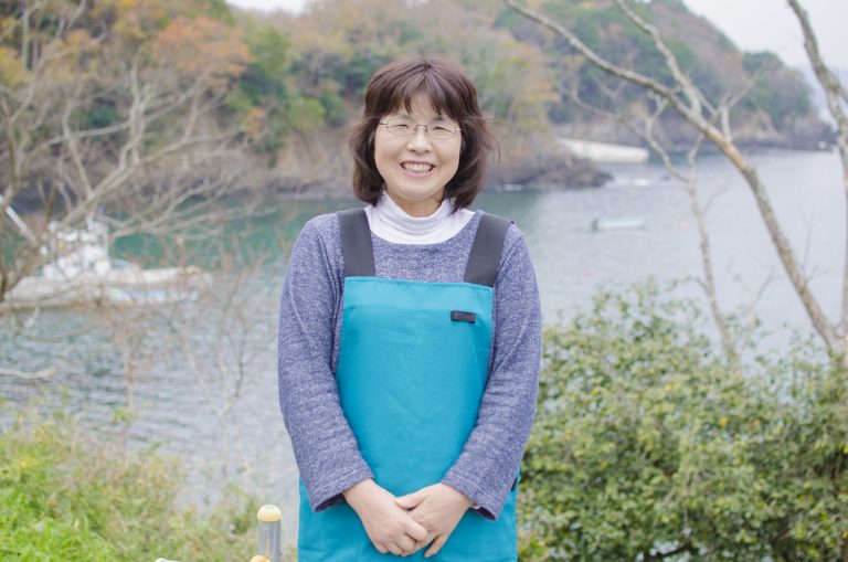 漁船が浮かんでいる海をバックに、水色のエプロンを着用した阿部さんが笑顔で立っている様子を撮影した写真