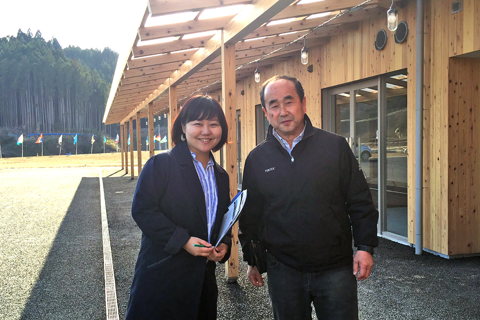 広い庇のある木造の建物の前に並んで立っている女性リポーターと高橋さんの写真