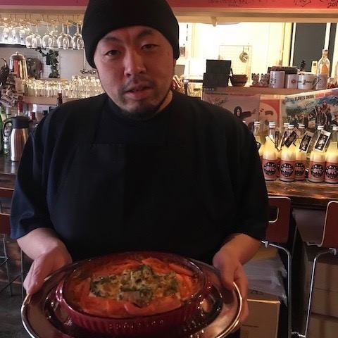 グラスや瓶入りの飲み物が並んだカウンターの前で、トマトなどが入った焼きカレーを乗せたお盆を持った内田さんが写っている写真