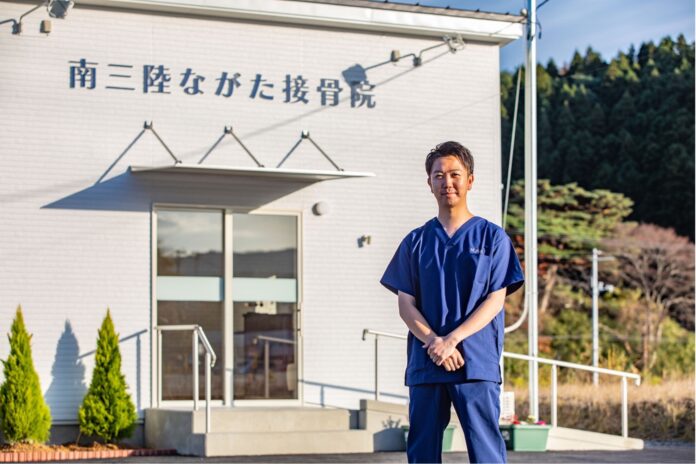 「南三陸ながた接骨院」と書かれた白い建物の前で長田滉平さんが青い施術着を着て笑顔で正面を向いて立っている写真
