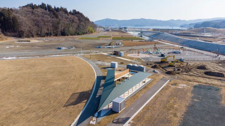 奥には森や山々が見え、周囲では造成工事が行われている広大な土地に、紙飛行機のような形をした屋根付きベンチと展望台が設置されているレンドリーステラスを上空から撮影した写真