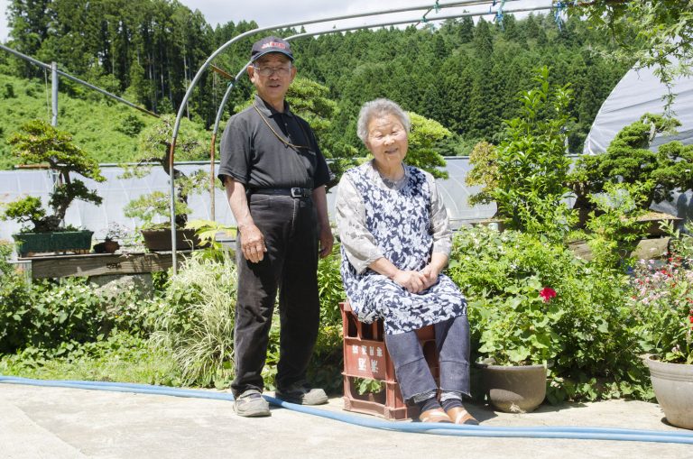 大きな盆栽や植物が育てられた庭の前に立つ黒色の服を着た男性と、逆さまに置いた赤色のビール瓶ケースの上に座った首藤さんの写真