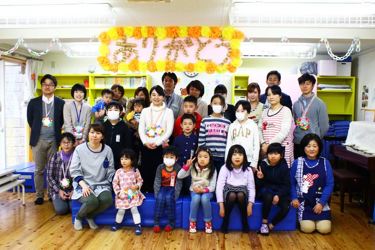 室内で「ありがとう」と書かれた花飾りの下に児童館の先生方と子どもたちが並び笑顔で記念撮影をしている写真