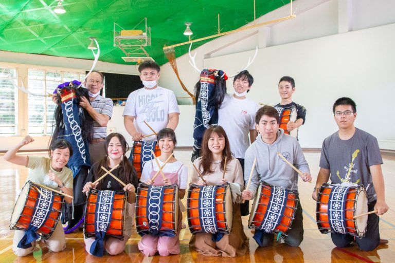 広々とした体育館で、太鼓を持った8人と、獅子頭を持った2人が2列に並んでいる写真