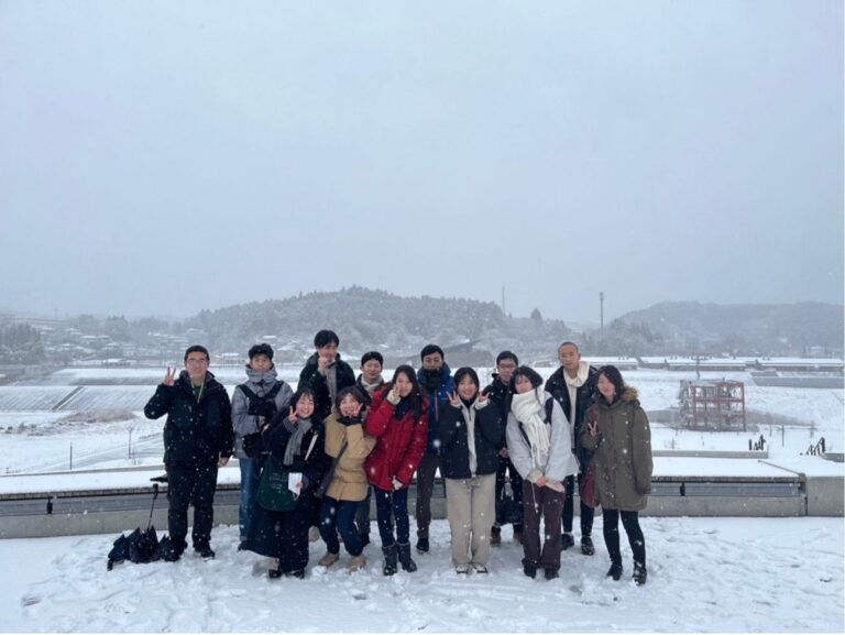 雪に覆われた山と建物が見え、雪が積もった広い場所で沢山の人々が並んでいる集合写真