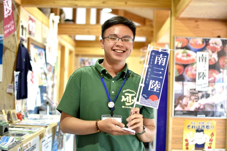 大きなポスターやシャツなどが飾られた室内に立つ、南三陸と書かれた小さな旗を両手で持つ笑顔の陳忠慶さんをアップで撮影した写真