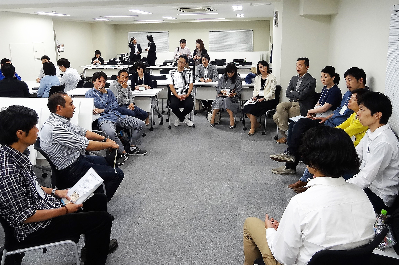 会議室でたくさんの男女が円型に置いた椅子に座り話し合いをしており、奥には別のグループが集まって話をしている様子が見える写真