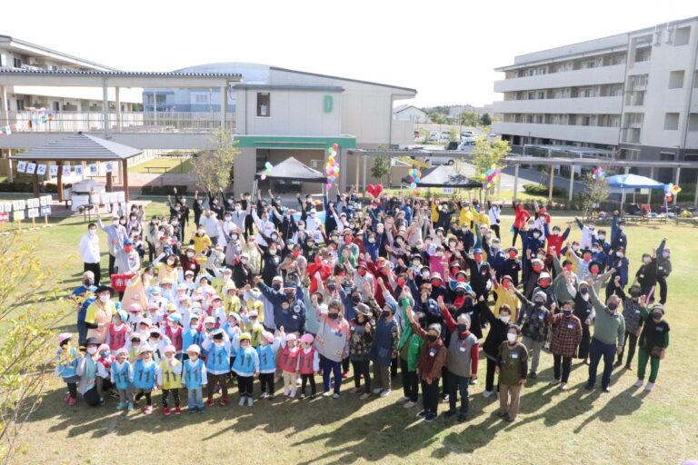 大勢の子どもや大人の参加者が屋外の広場に集まり、手を挙げて写っている「走らない“大運動会”」の集合写真