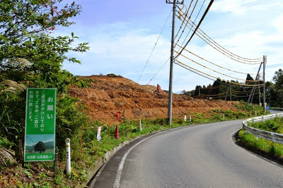 右にカーブする道路沿いの森が伐採され、造成が始まり土の斜面が見え、手前には「お願い」と書かれた緑の看板が立っている工事開始直後の写真