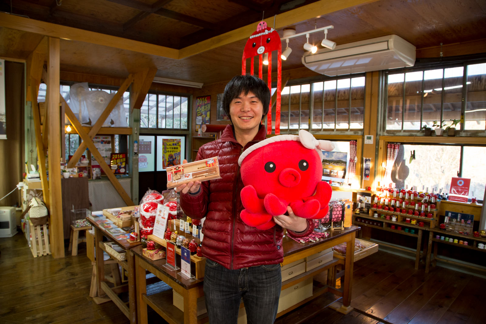 赤いタコのキャラクター「オクトパス君」のグッズが壁際や中央のテーブルにたくさん並べられた店内で、大きなオクトパス君のぬいぐるみとステーショナリーセットを持って立っている大森さんの写真