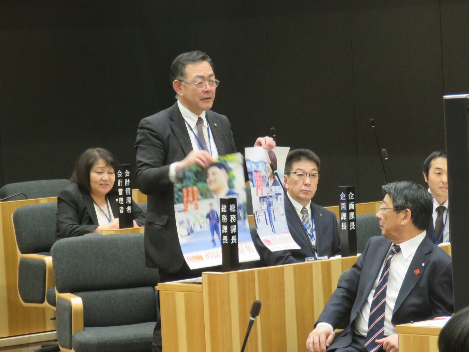 高橋総務課長が消防団員を募集するポスター2枚を持って立っている写真