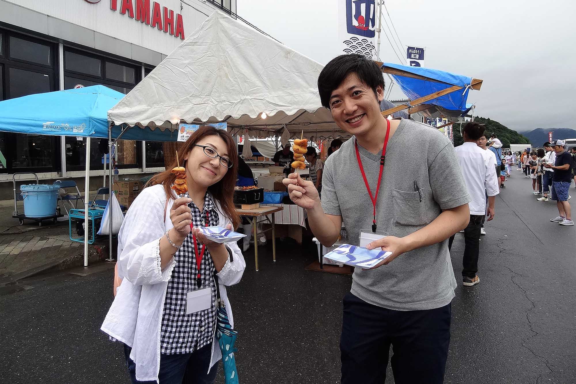 テント屋台の前で、女性と男性が笑顔でホヤの串焼を手に持っている写真
