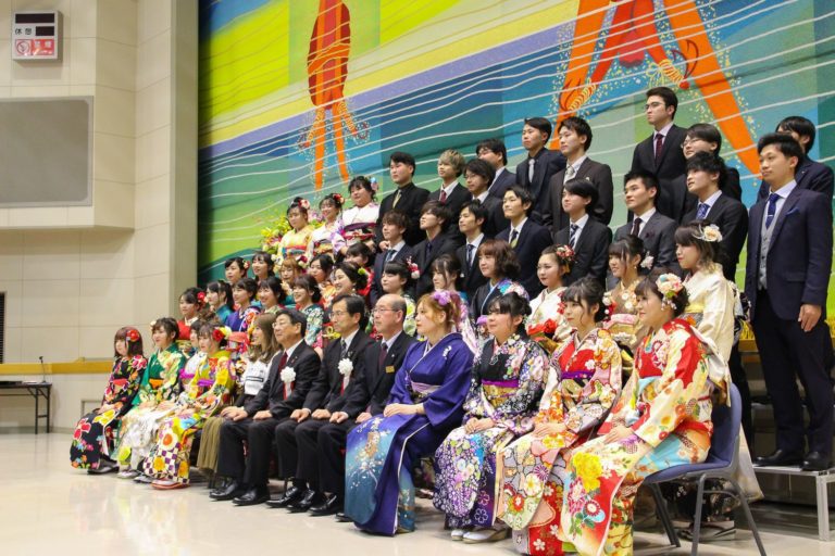 幕が下りたステージの下で、胸章リボンをつけた町長を中央にスーツ姿の男性や、艶やかな振り袖姿の女性たちが5列に並んでいる集合写真を右斜めから撮影した写真