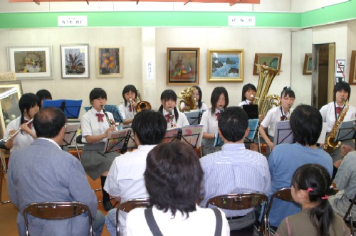 額縁に入った絵画が多数飾られたギャラリーの一室で吹奏楽部の生徒10人ほどが、楽器を演奏し、客席に座った来場者が鑑賞している様子の写真