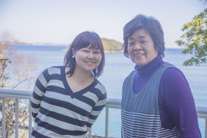 海を臨む高台でリポーターの女性と西條さんが笑顔で並んで立っている写真