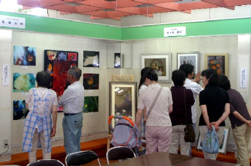 色々な絵画の作品が壁一面に展示された屋内のギャラリーで、複数の来場者がそれぞれの作品を熱心に鑑賞している、ミニ美術展の様子の写真