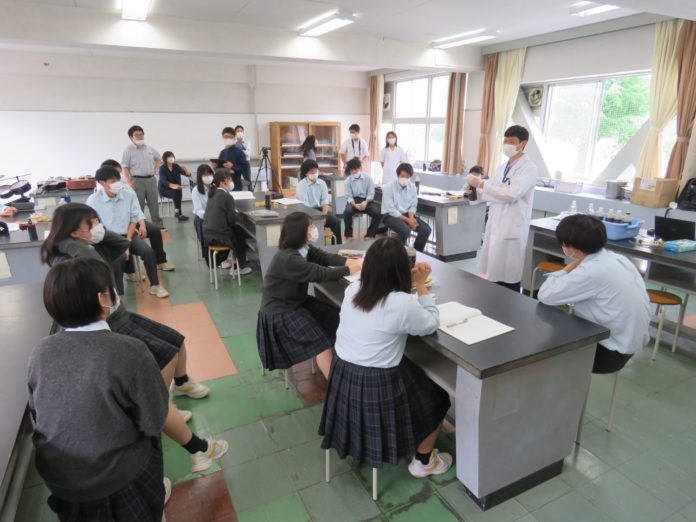 理科室のような教室で白衣を着たアミタ株式会社の職員の野添さんがマスク姿の学生たちに説明をしている様子の授業風景の写真