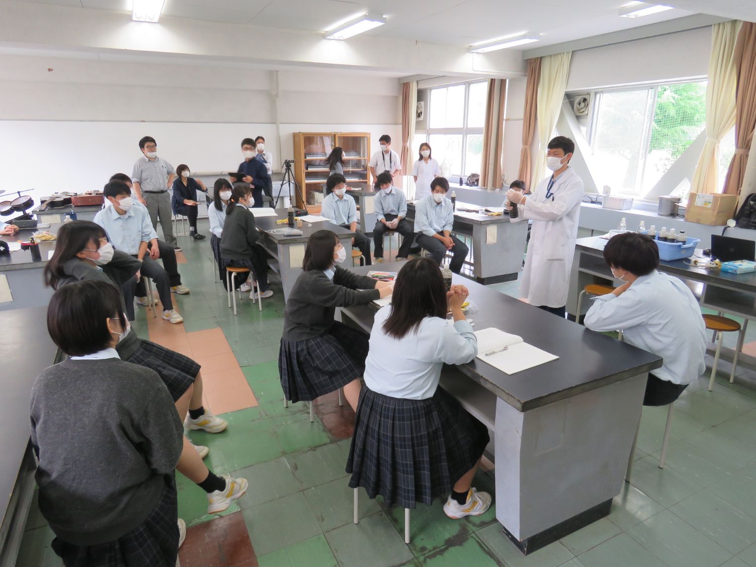 理科室のような教室で白衣を着たアミタ株式会社の職員の野添さんがマスク姿の学生たちに説明をしている様子の授業風景の写真