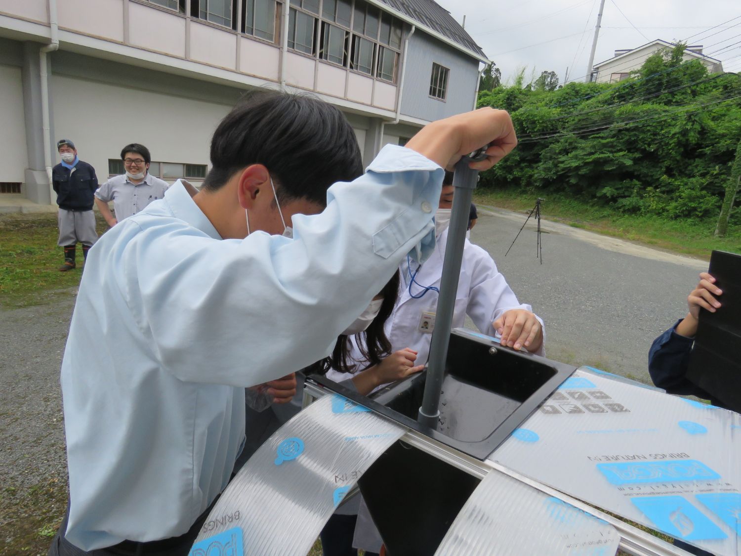 屋外で生徒が銀色のホームバイオガス装置の中を覗き込みながら実験作業を行っている様子の写真