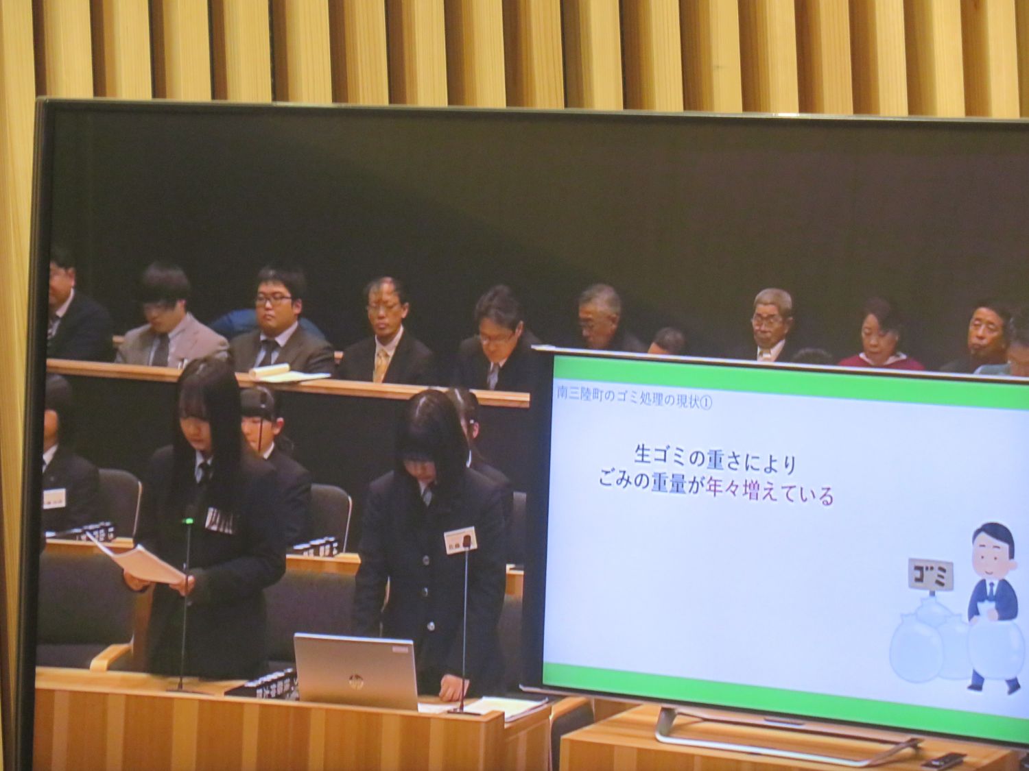 女子高校生たちが議場で「ごみ処理の現状」について発表し、スクリーンには「生ゴミの重さによりごみの重量が年々増えている」と書かれたスライドが映し出されている写真